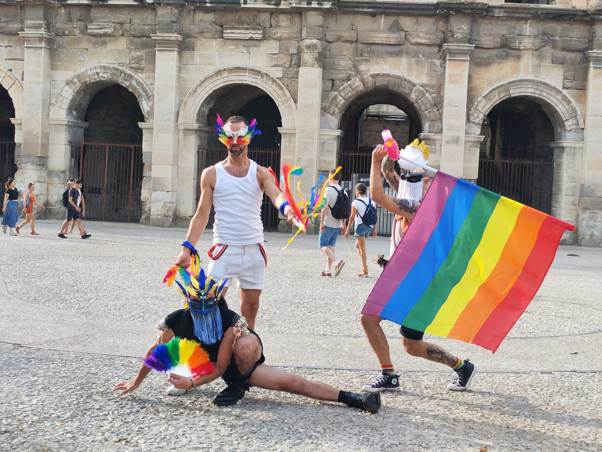 Personnes en costume avec drapeaux arc-en-ciel devant arènes.